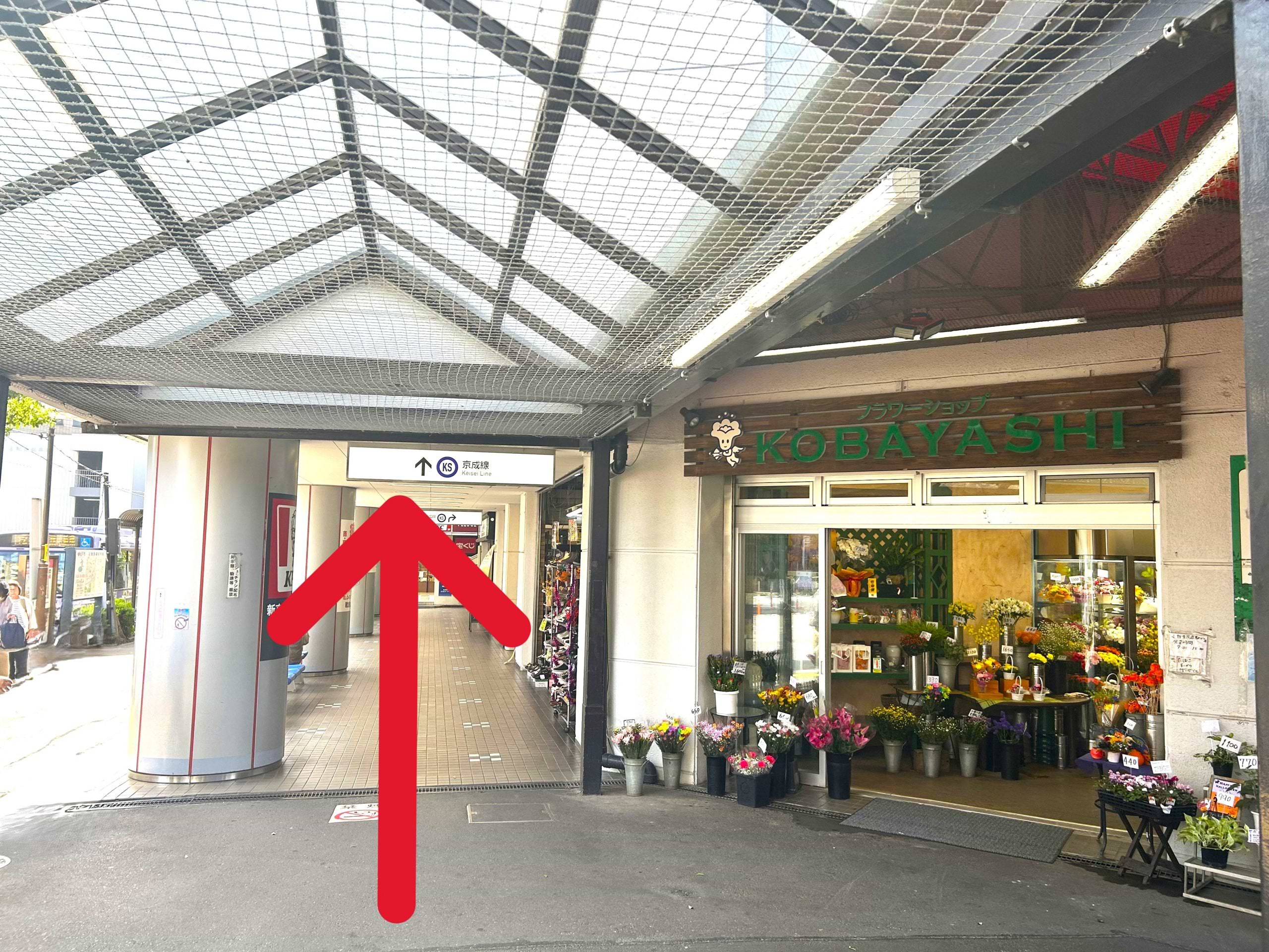 「小林生花店 京成駅ビル店」を右手に屋根付きの商店街（駅ビル）を直進します