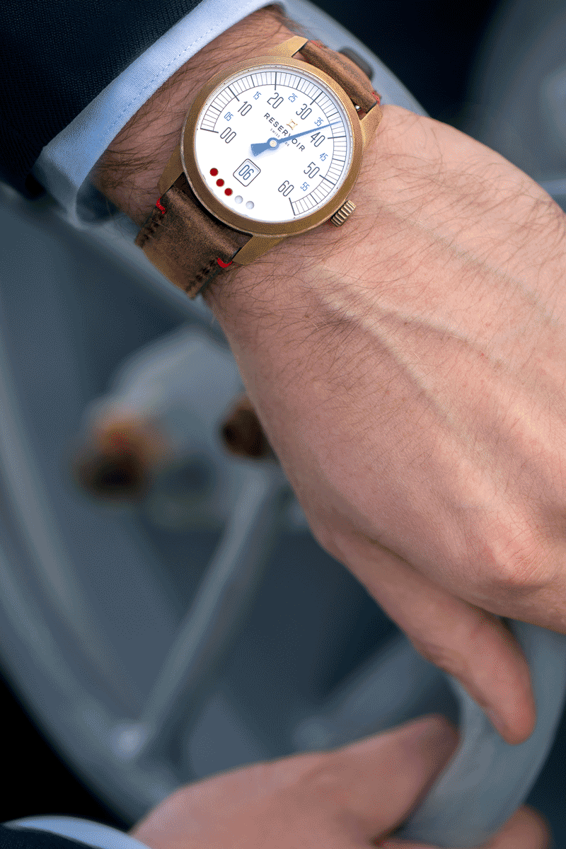 A wristwatch with a round white dial, bronze casing, brown leather strap on a wrist holding a lever.