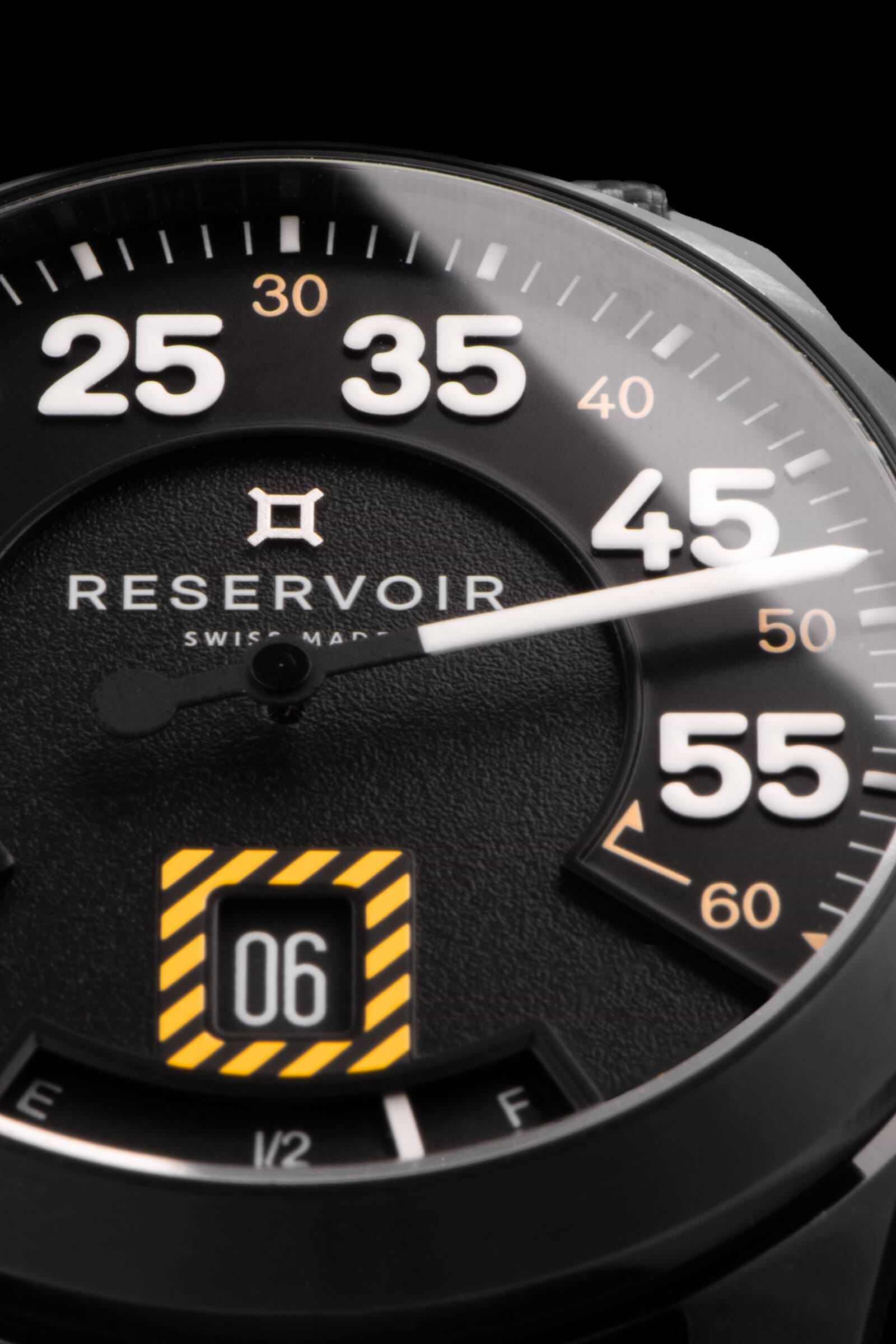 Gros plan du cadran d'une montre-bracelet Reservoir affichant des chiffres blancs, la date et un indicateur de type jauge à carburant.