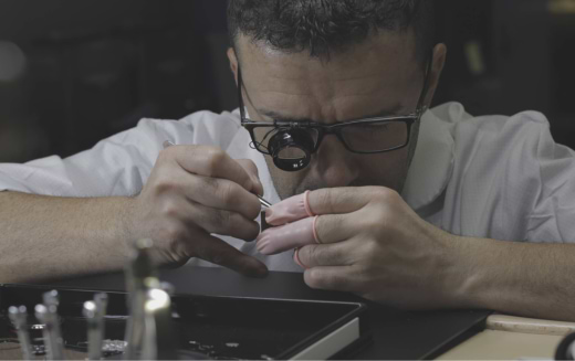 Person wearing magnifying glasses working on a small object with precision tools at a detailed workspace.