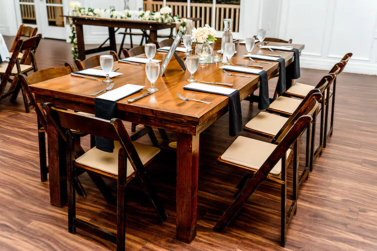 Elegant wedding reception setup featuring a beautifully arranged farm table with black napkins, glasses of water, and place settings. Chairs are arranged around the table, complementing the warm wooden tones of the flooring and decor.