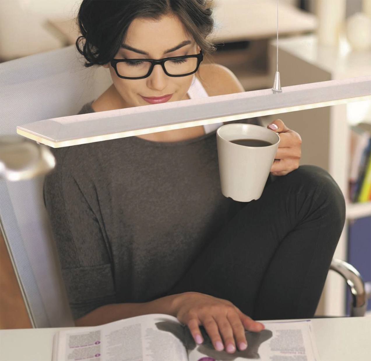 Hanglamp tafellamp Steinhauer Vrouw leest magazine aan tafel met kopje koffie in haar hand onder een Steinhauer hanglamp, wit en dimbaar.