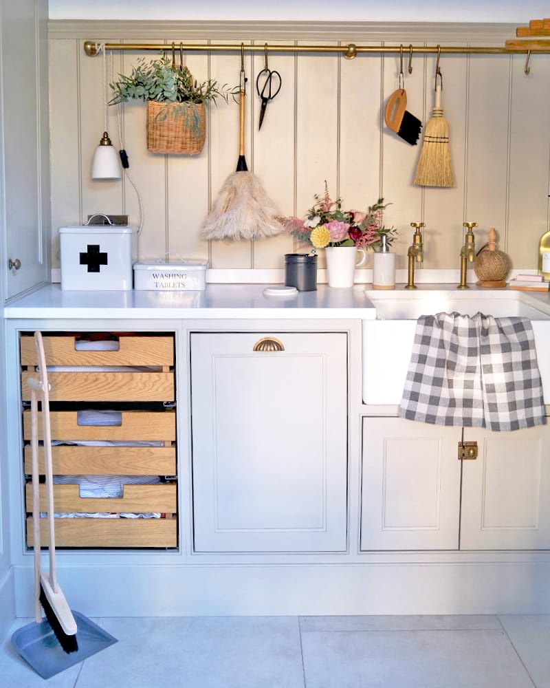 Utility room with grey in-frame cabinets accented by brass handles, surrounded by wood panelling. A white worktop holds a first aid box, washing tablet box, feather duster, garden scissors, brushes, an enamel jug, and a soap dispenser.
