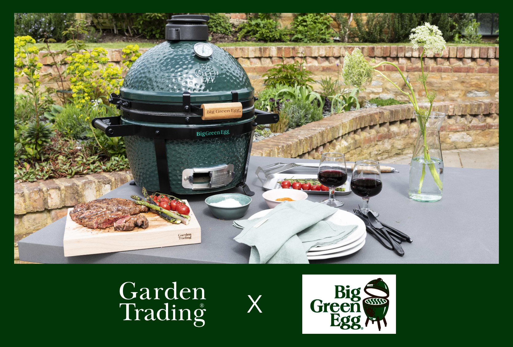 Big Green Egg MiniMax sitting on Garden Trading's Chilford Dining Table in a walled patio area, surrounded by Steak, Asparagus and Vine Tomatoes, plus grilling accessories