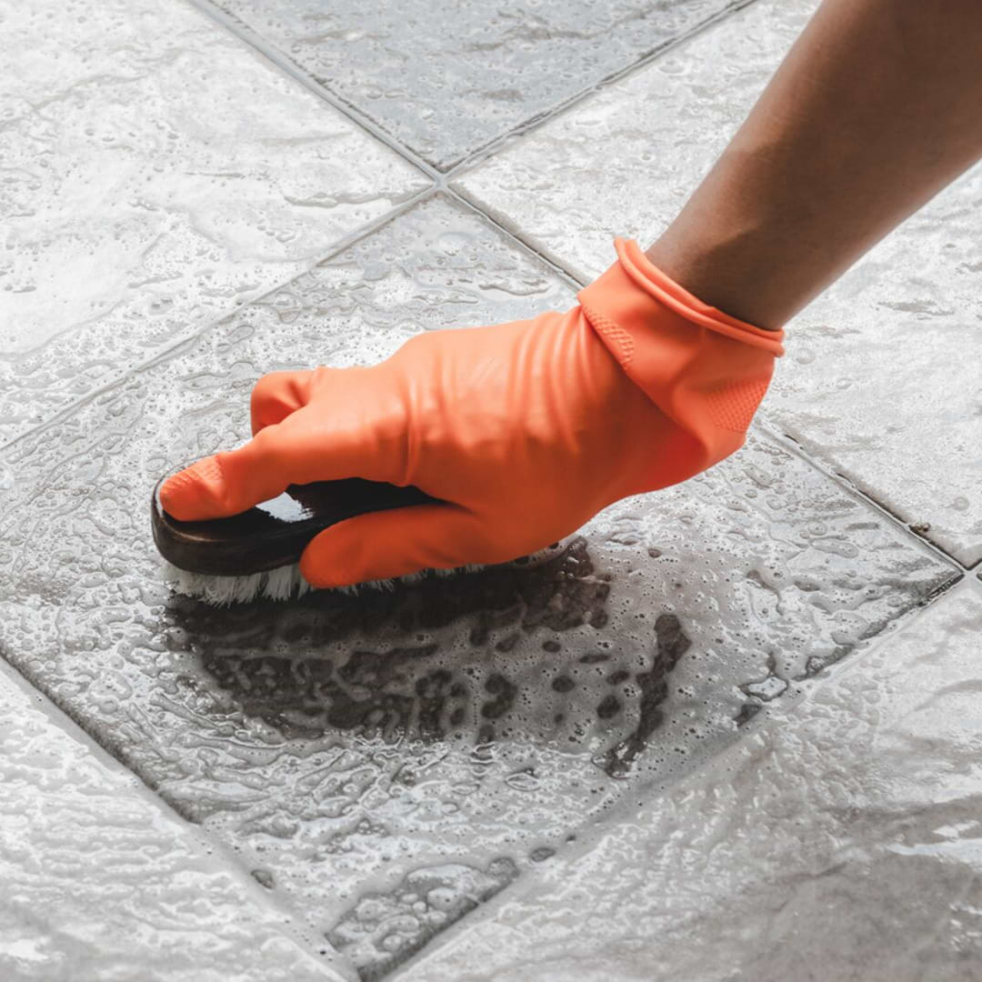 man's hand in orange glove scrubbing floor tile with brush - there are some bubbles and soap suds 