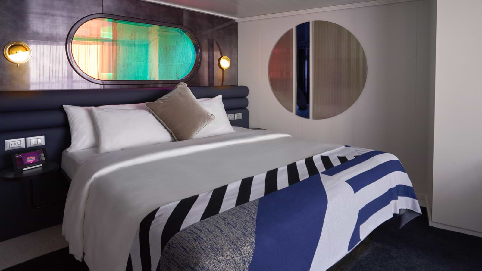 Virgin Voyages’ Seriously Suite view of bed with striped sheets, peek-a-boo shower window above and round mirror on the wall
