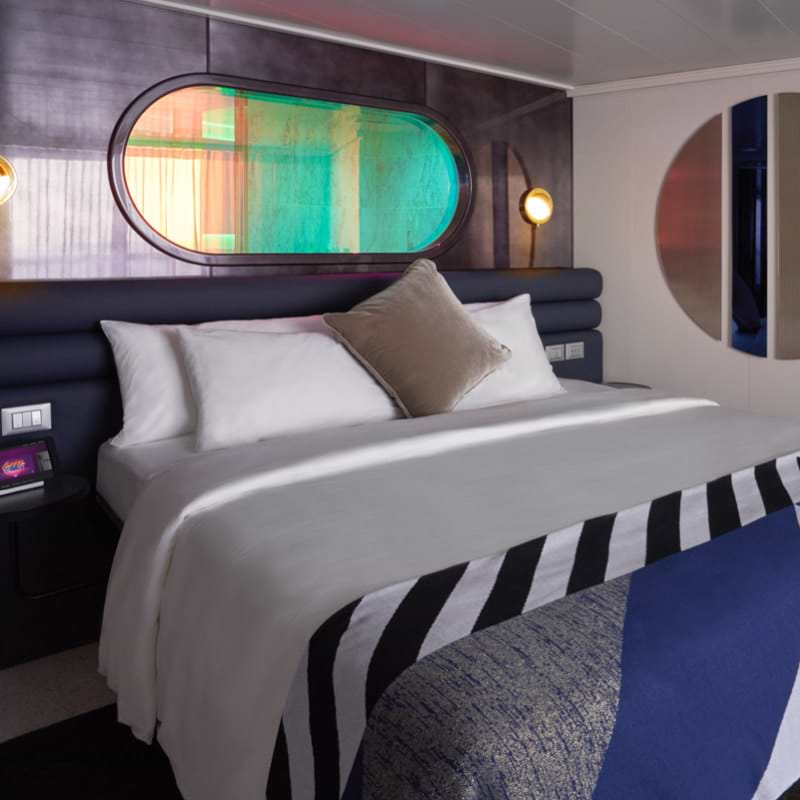Virgin Voyages’ Seriously Suite view of bed with striped sheets, peek-a-boo shower window above and round mirror on the wall
