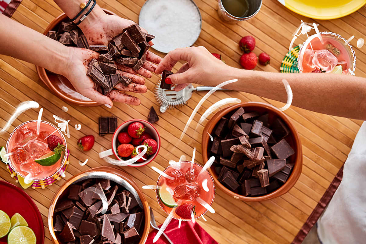Table with chocolate's bowls and Margaritas glasses, two hands offering chocolate to another hand and joyful graphics overlay