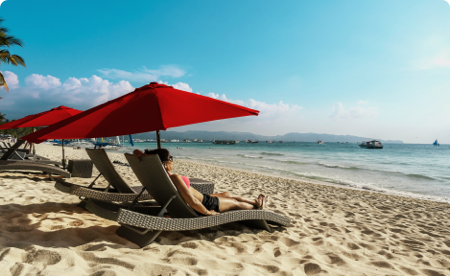Lounge chairs with beach umbrellas lined up in front of a calm beach, with a woman in sunglasses laying in the first chair.
