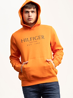 אדם עם קפוצ'ון אדום עשן של TOMMY HILFIGER עם לוגו מרכזי עומד על רקע בהיר רגיל, ידיים בכיסים.