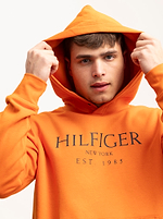 מישהו שלובש קפוצ'ון אדום עשן של TOMMY HILFIGER עם הלוגו של המותג בחזית מושך את מכסה המנוע מעל ראשו על רקע רגיל.